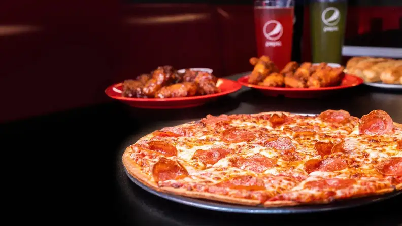 A pepperoni pizza in the foreground with wings and drinks in the background