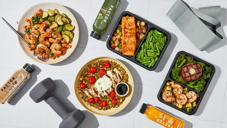 An overhead spread of Factor meals, including shrimp pasta, salmon with green beans, steak with broccoli, and Factor smoothies and juices, next to workout gear.
