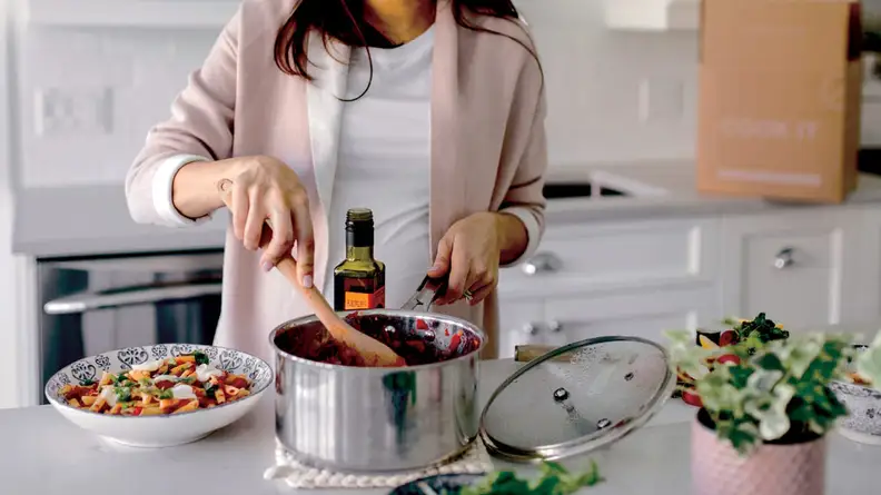 A woman preparing a meal in a stainless steel pot with the Cook it delivery box in the background.