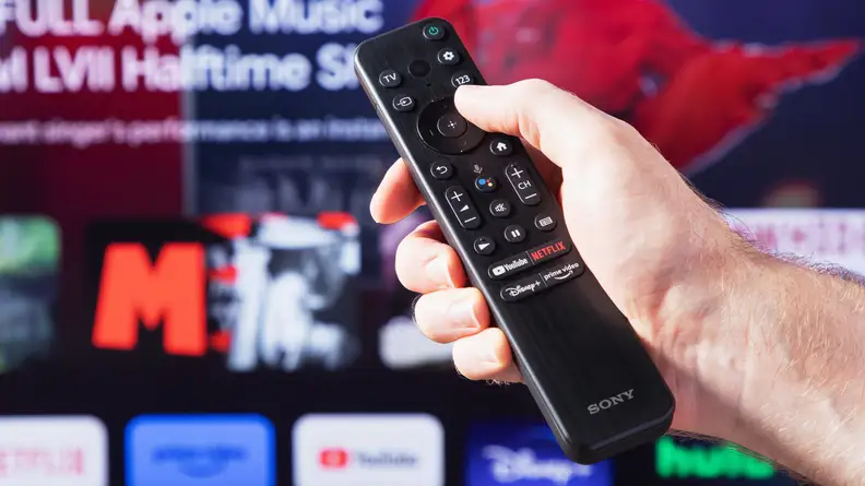 A person holding a Sony X90K LED TV remote with the Sony X90K LED TV's home screen, blurred in the background.