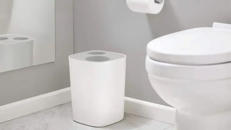 A all-gray bathroom contains a white toilet, a roll of toilet paper, and a trash can with an opening for trash and an opening for recycling.