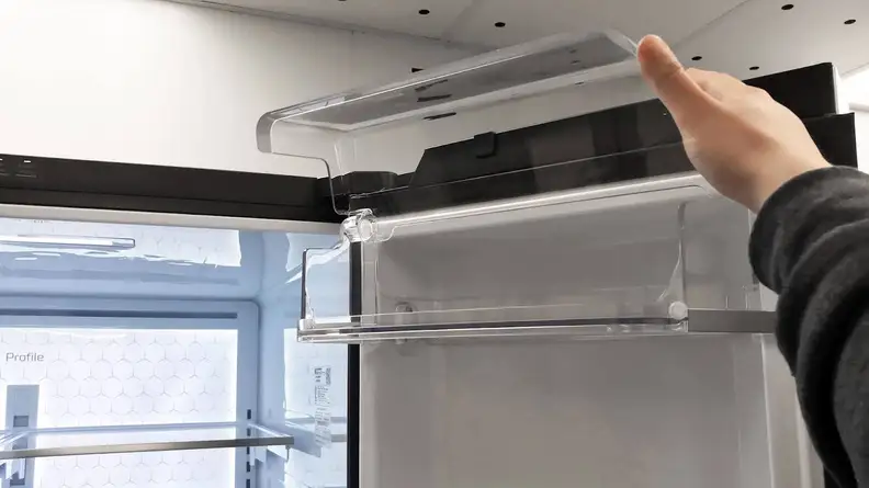 A hand reaches into frame, holding the dairy bin's lid open. The lid extends above the frame of the fridge.