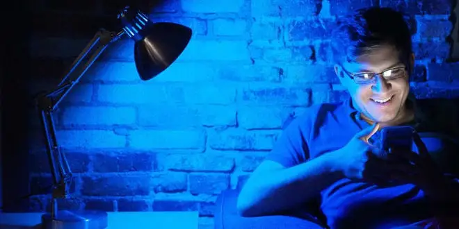 A man sitting on a couch in front of a brick background reading his phone, lit by blue light from a desk lamp.