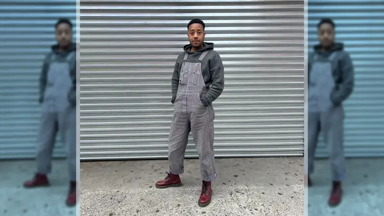 A person wearing overalls, red boots, and a gray hoodie, standing in front of a metal background.