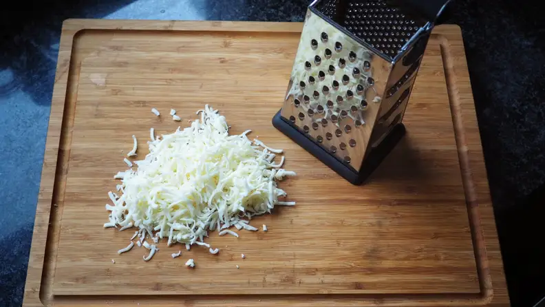 Some shredded cheese and a box grater on a wooden cutting board.