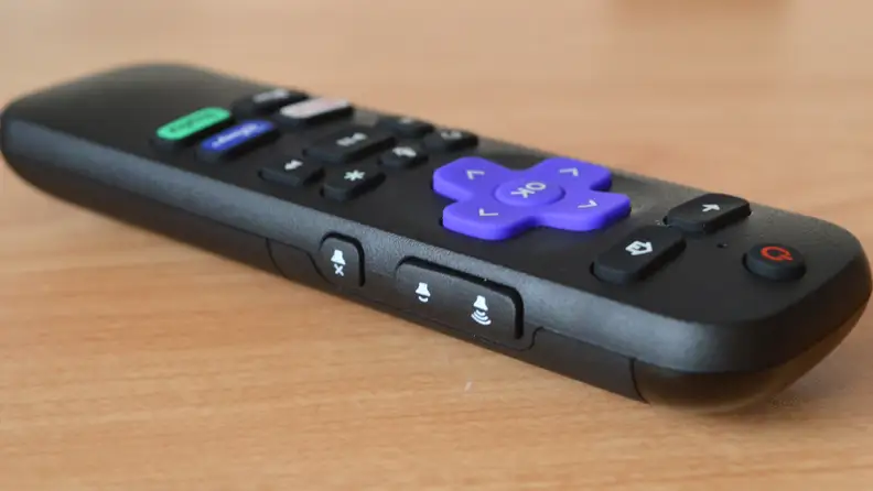 The Roku Express 4K+ remote is shown on its side, volume and mute keys out front. The remote sits on a woodgrain table and includes a purple navigation key and a power button.