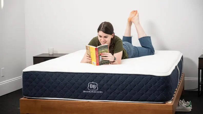 A woman lying on her stomach on a bare mattress, reading a book.