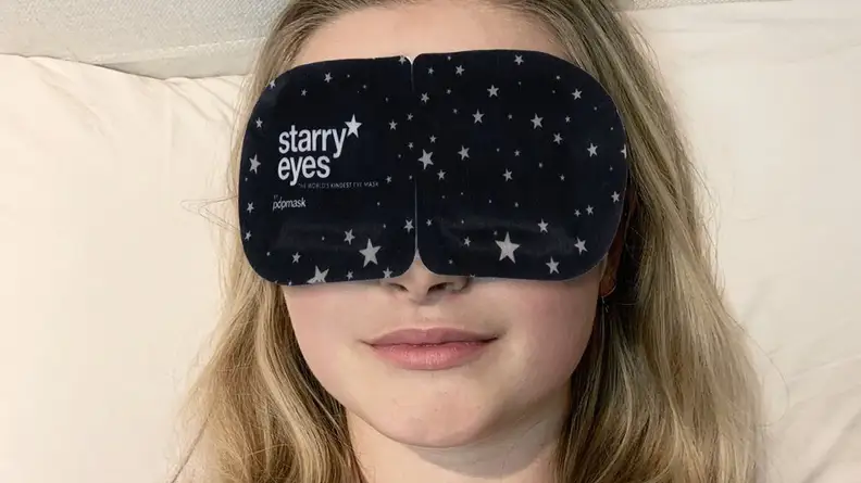 A person with long blonde hair wears a large black eyemask.