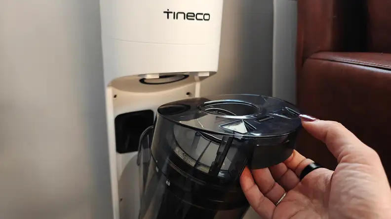 A hand removes a waste bin from a white Tineco vacuum base.