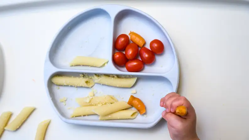 The Bumkins Silicone Grip Dish suction plate for kids with pasta and tomatoes in it, with a child's hand grasping food in the corner of the image.