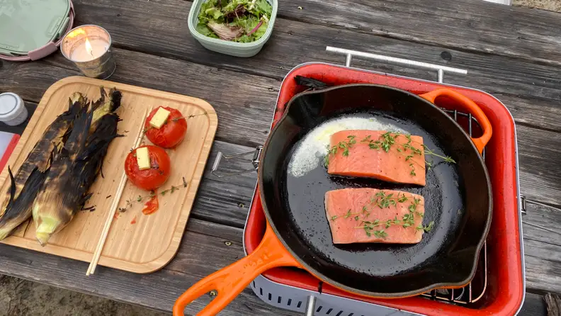 An orange cast iron skillet is placed on top of the Everdure Cube charcoal grill. In the skillet, there are two pieces of salmon. Next to the grill, there's a bamboo tray of cooked corn and tomatoes.