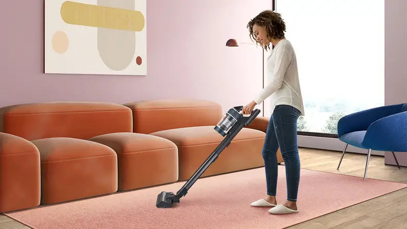 Woman cleaning a pink carpet with a cordless stick vacuum cleaner.