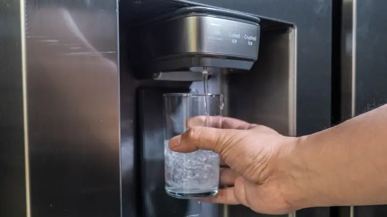 A hand reaches into frame holding a glass up to the Samsung RS27T5200SR's through-the-door dispenser.