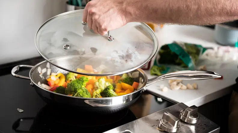 A person is lifting the lid off of a wok full of cooked vegetables.