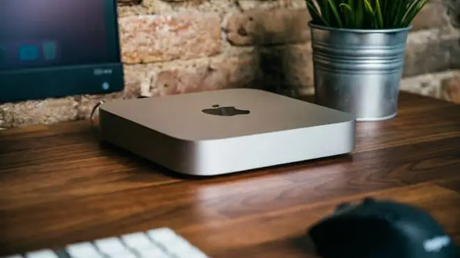 A silver Mac Mini sitting on a desk with a mouse, a travel mug, a keyboard, and part of a monitor.