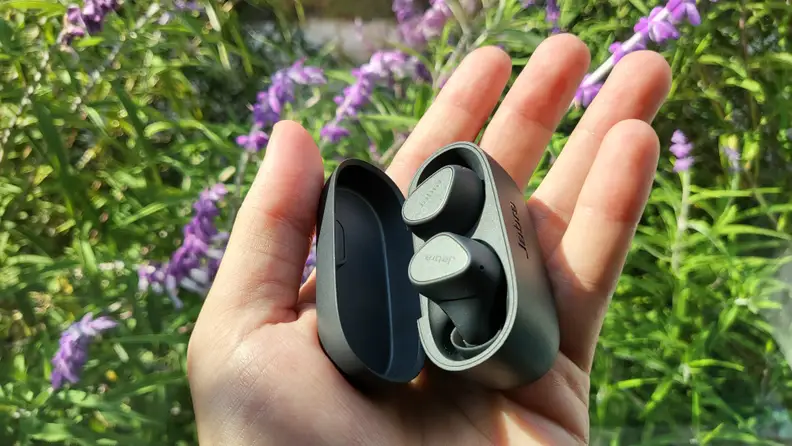 Jabra's all-black, matte earbuds sit in their open case in an open hand above green and purple flowers blown in a light breeze.