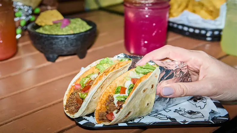 A hand holding a plate of tacos, with a margarita and guacamole in the background