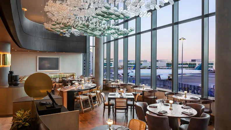 Upscale airport restaurant with floor-to-ceiling windows overlooking planes on the runway, featuring modern décor, candlelit tables, and a sculptural ceiling installation.
