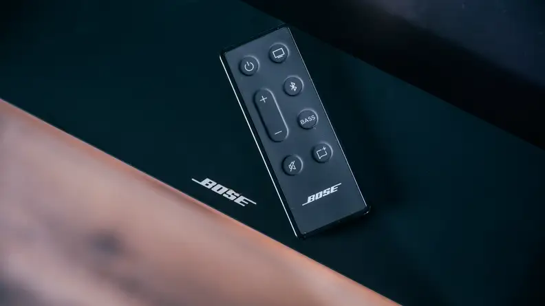 The Bose TV Speaker remote resting on top of the all-black soundbar, the white Bose logo peaking out beneath the slim rectangular remote with bubbled buttons on top.