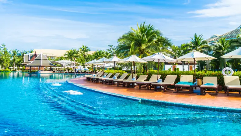 A luxurious resort pool with lounge chairs, umbrellas, and palm trees under a bright blue sky.