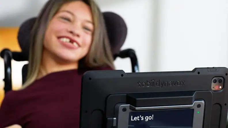 A little girl in a wheelchair looking at an AAC device.