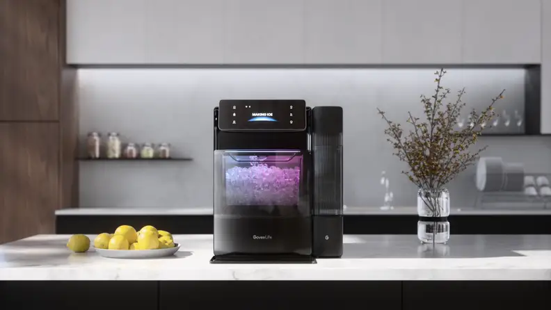 A countertop ice maker sits in a modern kitchen, with a clear window showing freshly made ice illuminated inside the machine.
