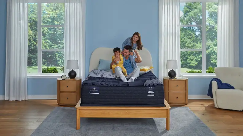 A father and two children playing on a Serta Perfect Sleeper X hybrid mattress in a bright bedroom.