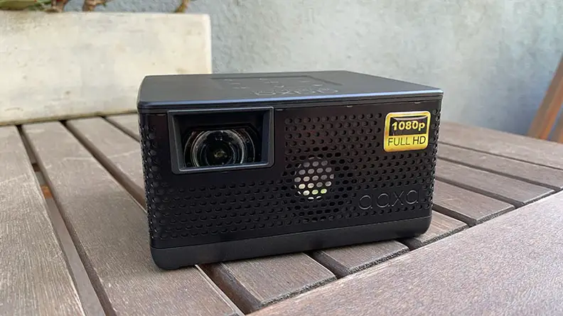 The AAXA P400 portable projector sitting on a wooden table outdoors in front of a stucco wall.
