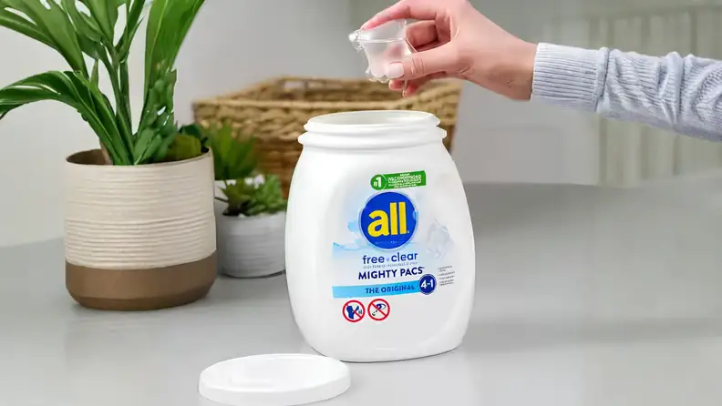 A hand drops an All Free Clear Mighty Pacs laundry pod into an open tub on a countertop next to potted plants.