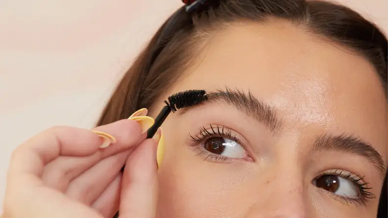 A closeup on the top half of a model's face as they hold a spoolie up to their eyebrow.