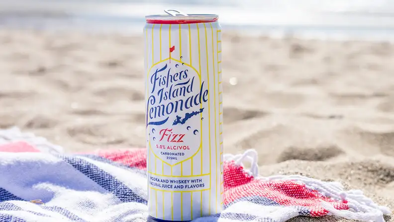 A can of spiked lemonade sits atop a beach towel on a sandy beach.