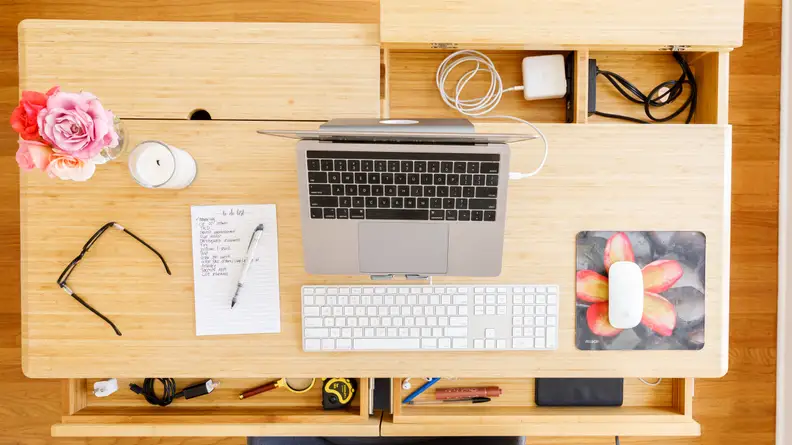 The top of a desk showing a computer, a pair of glasses, and a vase of flowers