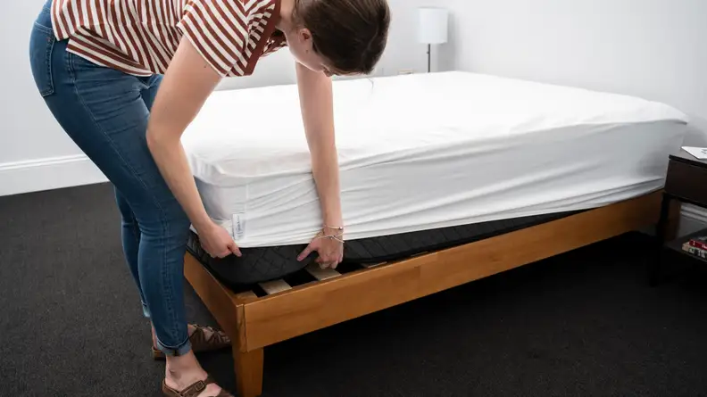 A person pulls a mattress protector over the edge of a bare mattress