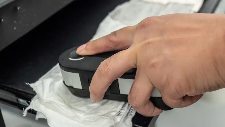 A hand reaches into frame holding a photospectrometer up to a washed stain strip, measuring exactly how much stain was scrubbed away.