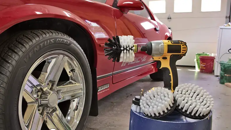 Cordless drill sitting on top of bucket next to brush attachment pieces adjacent to red sports car.