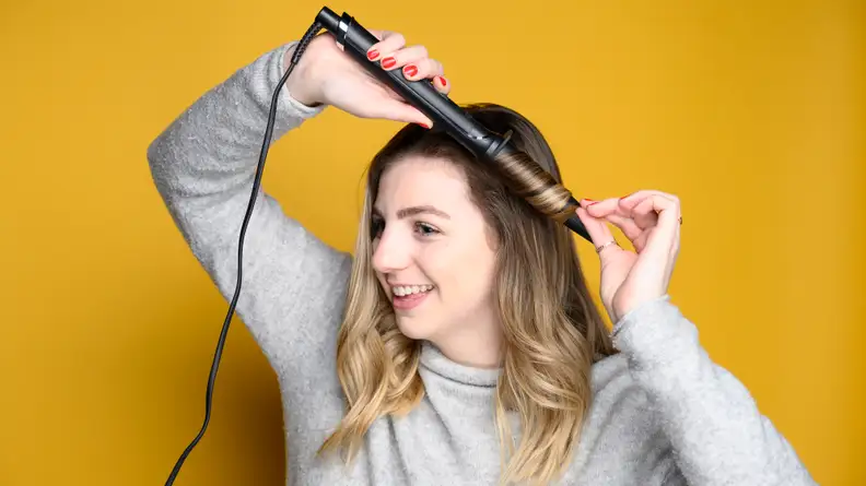 A person with long hair holds a curling iron in their hair and smiles.