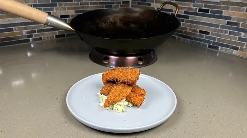 Small fried fish pieces on top of small gray plate in front of wok cooking pan.