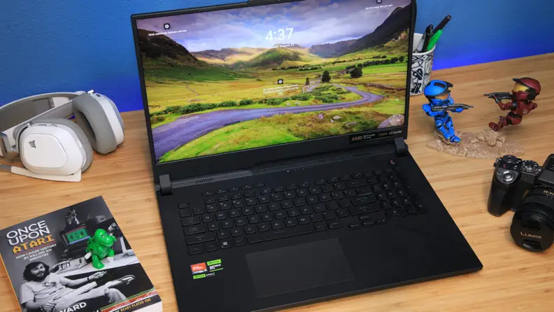 Asus ROG Strix SCAR 17 gaming laptop sitting on desk surface next to books, a camera, a pair of headphones and cup of pens.
