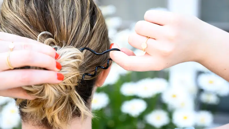 The back of a woman'd head as she pulls her blond hair into a messy bun with a coil hair tie.