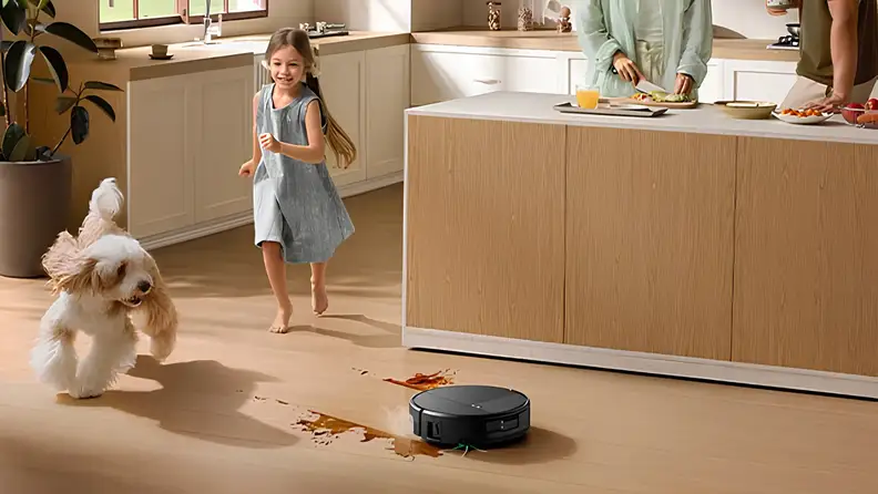 A young girl runs through a kitchen with a fluffy white dog while parents cook at the island, and an iRobot Roomba robot vacuum mops a sauce spill on the hardwood floor.
