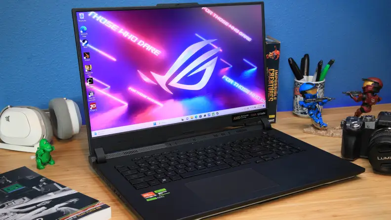 Asus ROG Strix SCAR 17 gaming laptop sitting on desk surface next to books, a camera, a pair of headphones and cup of pens.