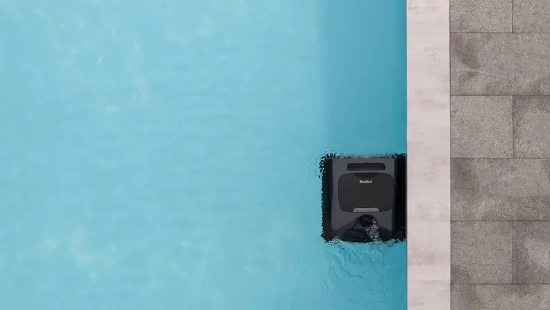 A black, square robot sits on the surface of the water by the edge of a swimming pool