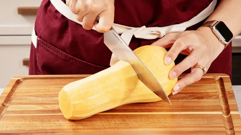 Person using knife to cut butternut squash in half horizontally on top of wooden cutting board.