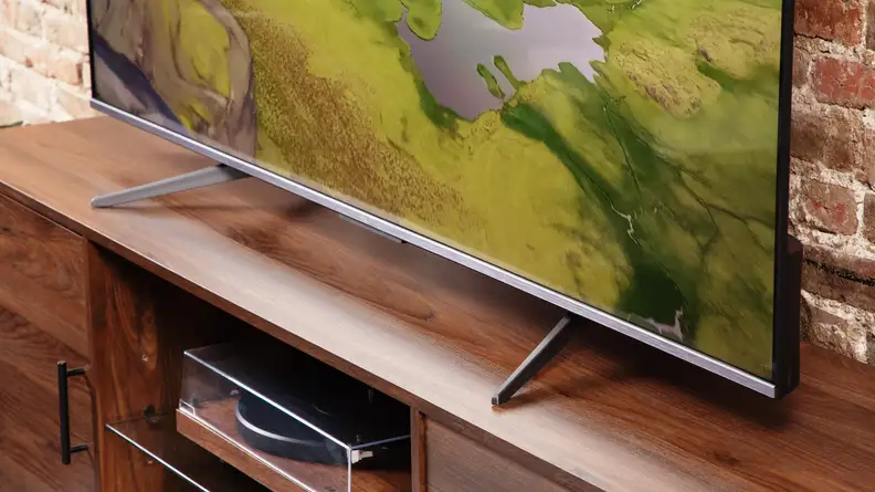 TCL Q6 LED TV on top of wooden stand in front of brick wall with green mountains on screen.