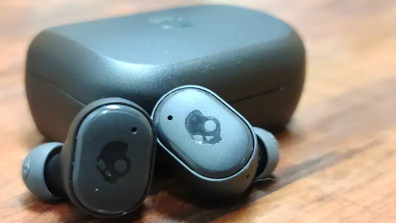 The all-black Skullcandy Grind Fuel sit in front of their black case on a wooden table with the eartips pointed in and a skull carved into the exteriors.