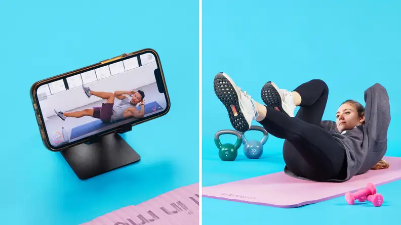 On left, smart phone perched on phone stand with FitOn app on screen. On right, Person laying on yoga mat with feet elevated in the air and arms behind head completing a workout.