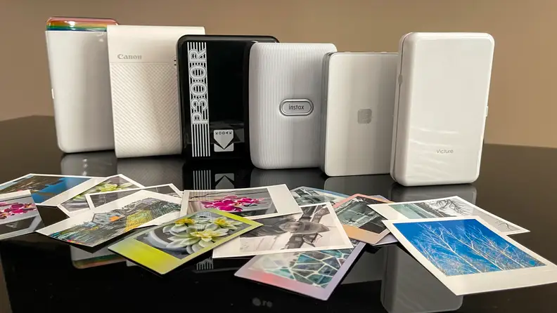 The Polaroid Hi-Print, Canon Ivy, Kodak Step, Fujifilm Instax Mini Link, Lifeprint 2x3 Hyperphoto, and Victure PT320 portable photo printers lined up on a table with a collection of photos in front of them.