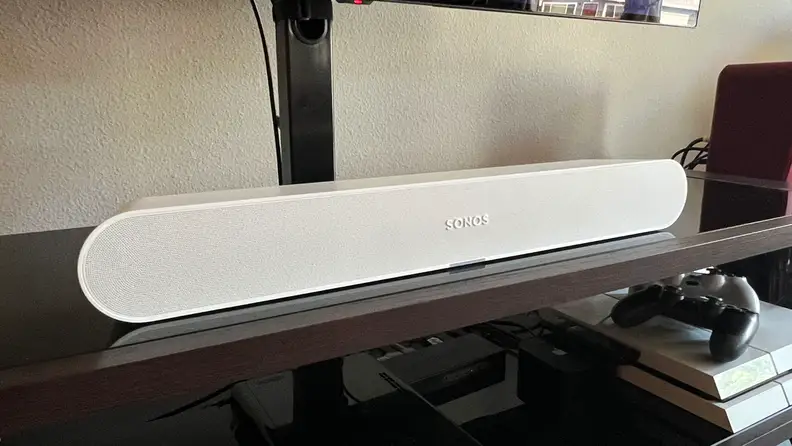 The white Ray soundbar is shown above a PS4 console on a black and brown TV console.