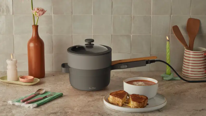 The Perfect Power Pot in char color sitting on a countertop next to a vase, candles, cutlery, and a plate with a sandwich and soup.