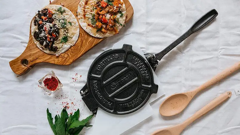 Product shot of Victoria cast iron tortilla press next to tortillas, spices and wooden spoons.
